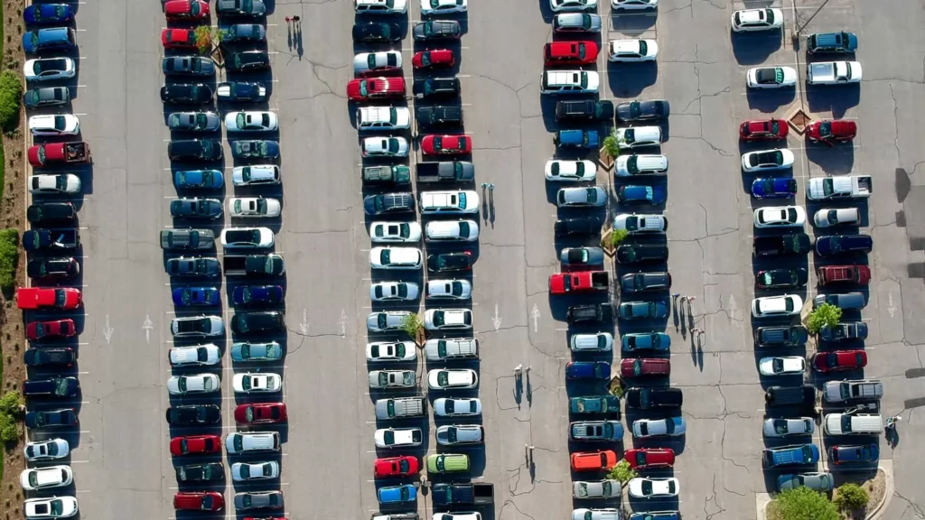 Aerial view of a large parking lot with rows of parked cars, including some electric cars at EV charge stations, and people walking between the rows on a sunny day. The pavement shows visible cracks and lines separating parking spaces.