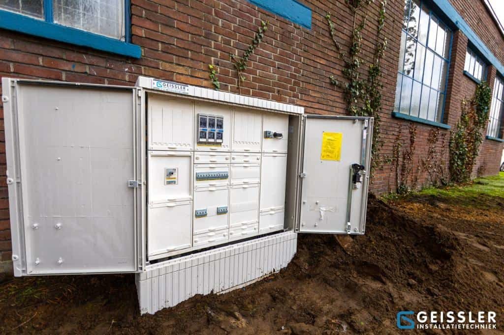 An outdoor electrical control panel with open doors is installed against a brick building. The dug-up ground suggests recent work on charge connections, and the panel displays meters, switches, labels, and the Geissler logo.