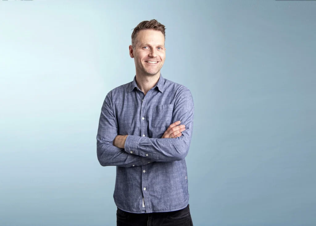 A smiling man with short brown hair stands with arms crossed, wearing a blue button-up shirt and dark pants, against a plain light blue background, as if ready to discuss EV charging or the future of electric cars.