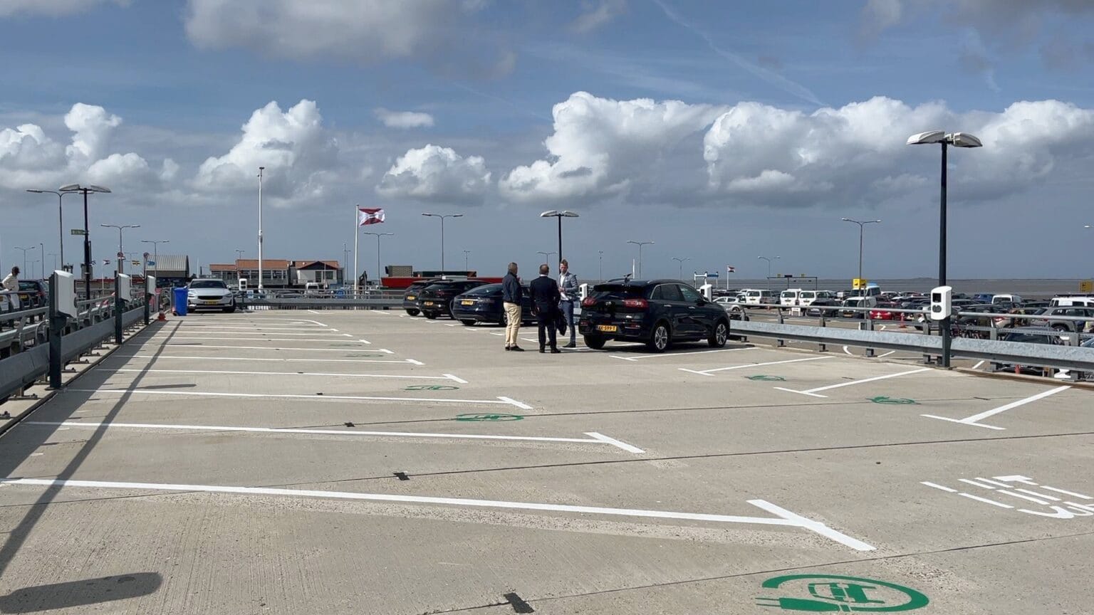 A mostly empty rooftop parking lot under a partly cloudy sky, with three people standing near parked electric cars. Several open charging spaces are visible, and distant buildings and cars can be seen in the background.