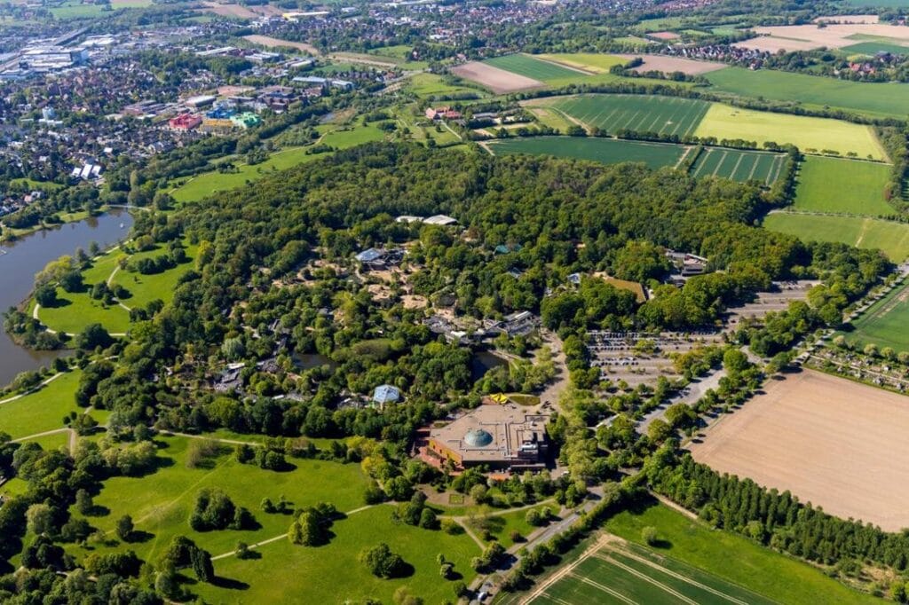 Aerial view of a large park with dense trees, walking paths, a lake, open fields, several buildings with EV charging stations, parking lots, surrounded by farmland and a distant town.