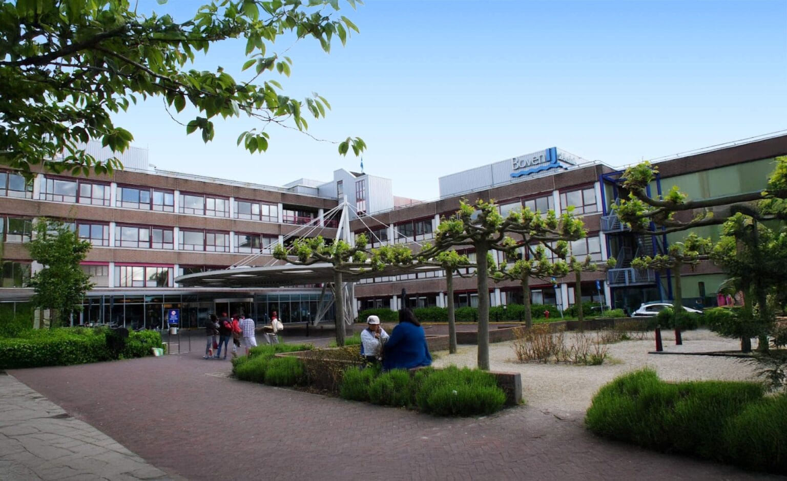 People sit and walk in a landscaped courtyard in front of a modern, multi-story building with large windows and a sign that reads Hoven. Free EV charging stations are available near the entrance, surrounded by trees under a clear blue sky.