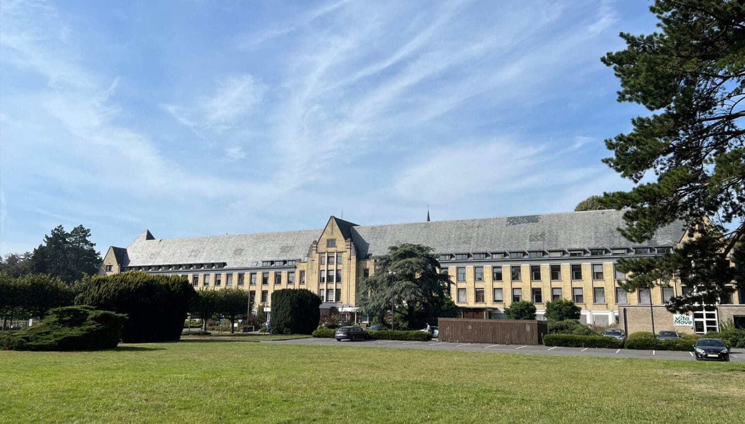 A large, yellow-brick building with a slate roof and multiple gables stands behind a green lawn and parking area featuring Pluq free loading stations for charging cars. Trees and bushes are scattered around under a blue sky with wispy clouds.