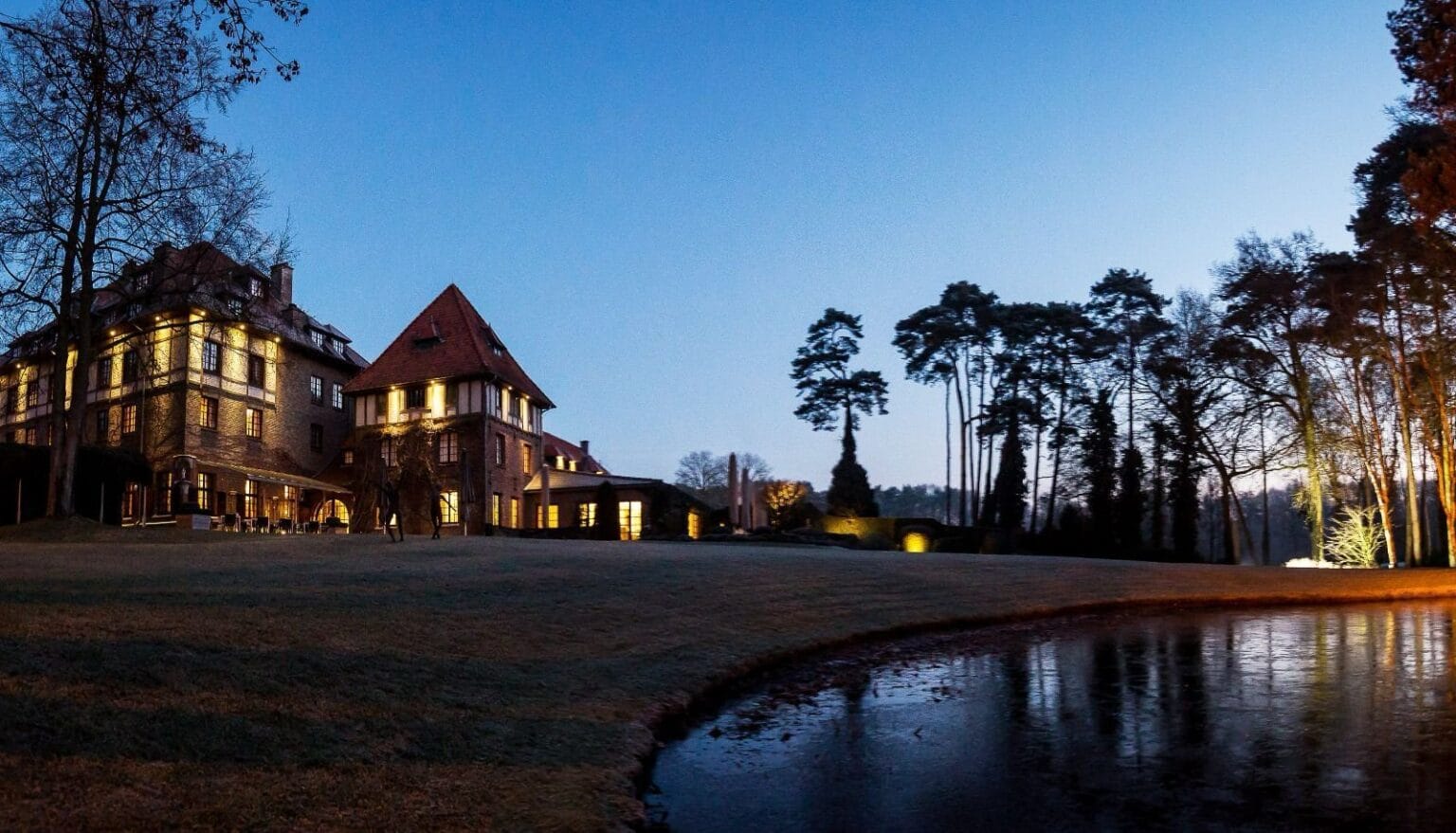 A large, elegant brick mansion with lit windows stands beside a pond and lawn at dusk, surrounded by tall trees and featuring free loading stations for electric cars against a clear evening sky.