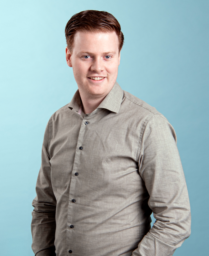 A man with short, neatly styled brown hair is wearing a light gray button-up shirt and smiling at the camera, standing against a light blue background near free loading stations for electric cars.
