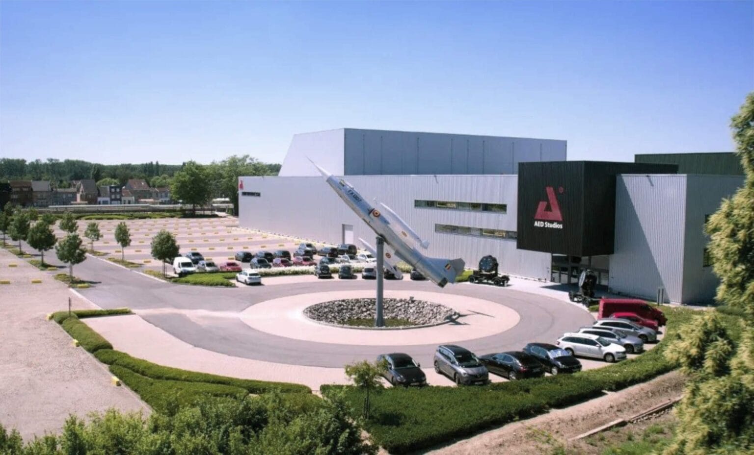 A large white industrial building with MSD Studios signage, surrounded by parked cars and trees. In front, a model rocket stands upright in a circular driveway featuring free loading stations for electric cars. A parking lot and small buildings are visible in the background.