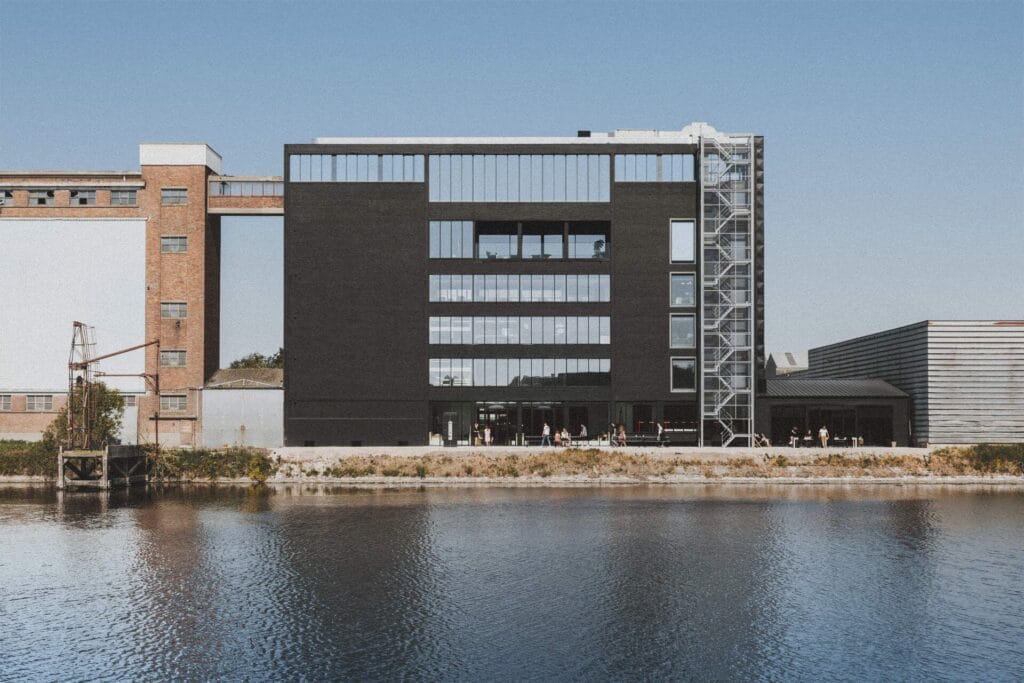 A modern black building with large windows stands by the water, featuring an external metal staircase on the right. Near the entrance, people gather by charging stations from pluq, while older industrial buildings are in the background.