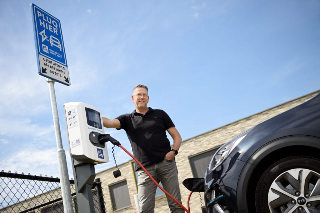 A man stands next to an EV charging station, holding the cable plugged into a black car, with a blue sky and building behind him. A sign above marks it as a free loading station for EVs to charge.