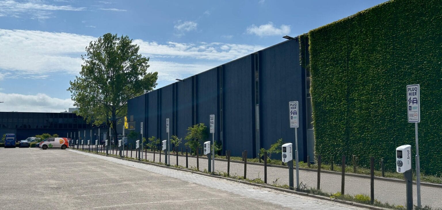 An empty parking lot features several EV charging stations and signs reading PURE along a building partly covered in greenery, all beneath a blue sky with scattered clouds.