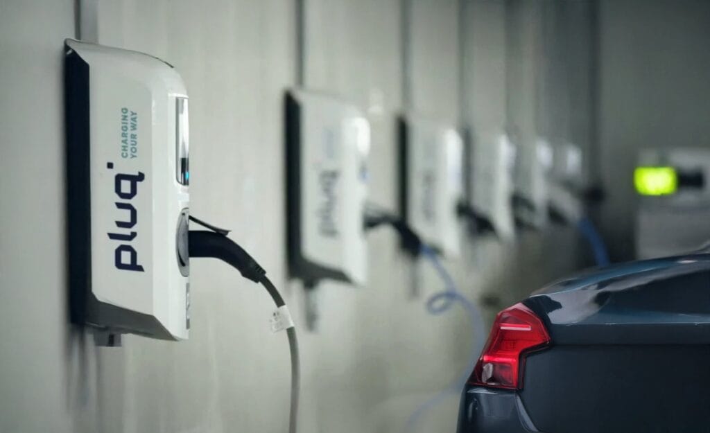 A row of electric vehicle charging stations is mounted on a wall, with one dark-colored car charging. The pluq stations offer convenient free loading stations in an indoor parking facility.