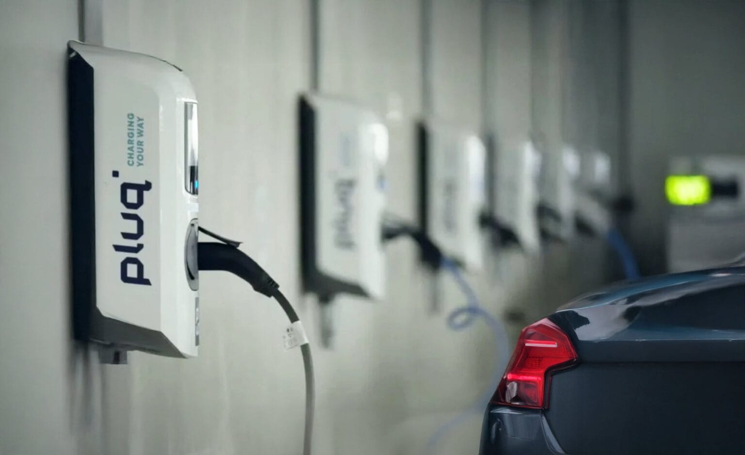 A row of electric vehicle charging stations is mounted on a wall, with one dark-colored car charging. The pluq stations offer convenient free loading stations in an indoor parking facility.