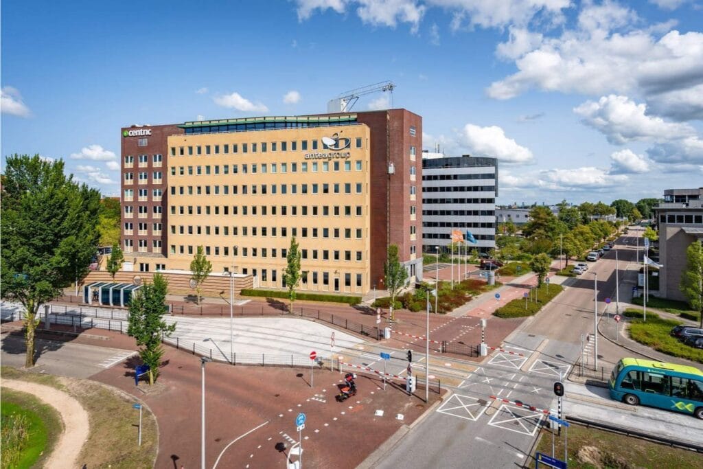 A large, multi-story office building with Amersfoort signage, located on a corner with surrounding trees, roads, bike lanes, and a few people and cars nearby—plus free loading stations for charging under a partly cloudy sky.