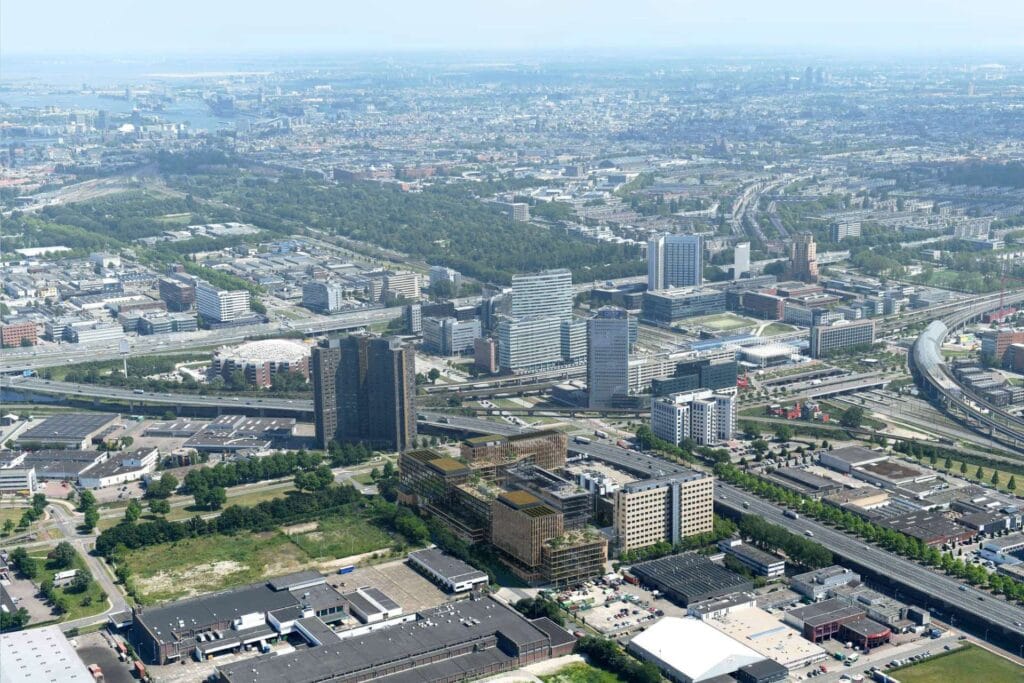 Aerial view of a cityscape with modern office buildings, green spaces, and roads featuring EV charging stations, highways stretching across the wide city expanse, and water visible in the distance under a clear sky.