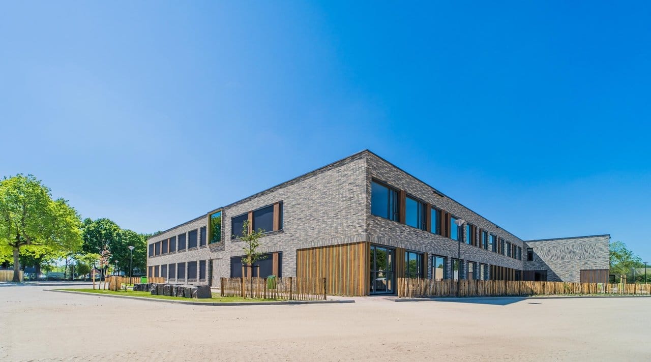 A modern, two-story building with a gray brick exterior and large windows, surrounded by a wooden fence and trees under a clear blue sky, features free loading stations for electric cars to charge.
