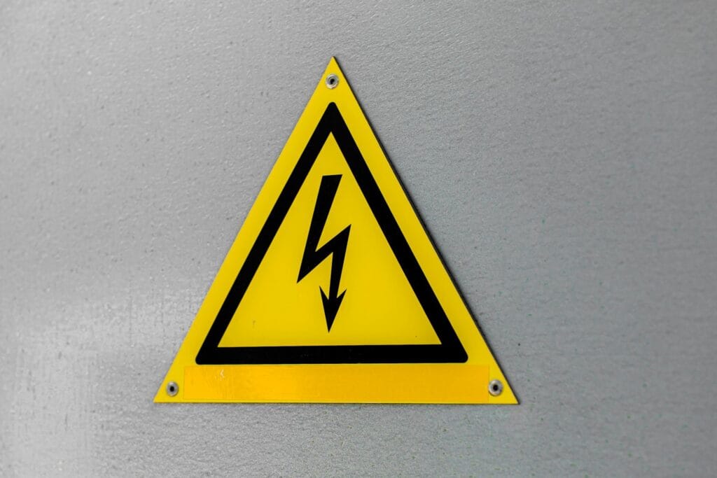 A yellow triangular warning sign with a black border and a black lightning bolt symbol, indicating a high voltage or electrical hazard, mounted on a gray wall.