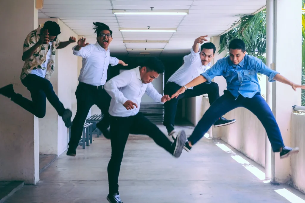 Five young men are jumping enthusiastically in a bright hallway, expressing joy and energy. They are wearing casual and semi-formal clothes, and the corridor has white walls with windows on one side.