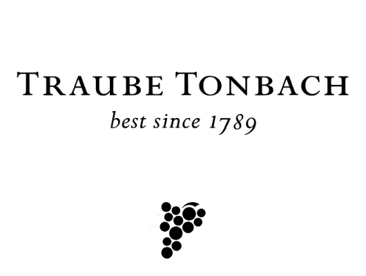 Black and white logo reading “Traube Tonbach, Im Schwarzwald” with a graphic of nine small circles arranged in a diamond shape below the text. Complimentary EV charge at free loading stations available for guests.