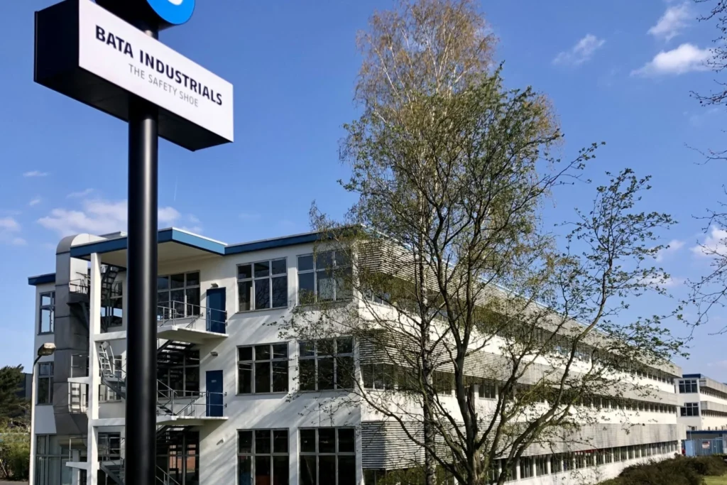A modern white building with large windows and blue accents sits behind a tree. In the foreground, a tall sign reads Bata Industrials The Safety Shoe, hinting at innovation—perhaps even exploring charging as a service solutions. The sky is clear and blue.