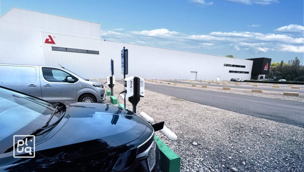 Two electric vehicles are parked at charging stations outside a large white industrial building with red triangle logos, on a sunny day with blue sky and clouds. The foreground shows “plug” branding on a black car.