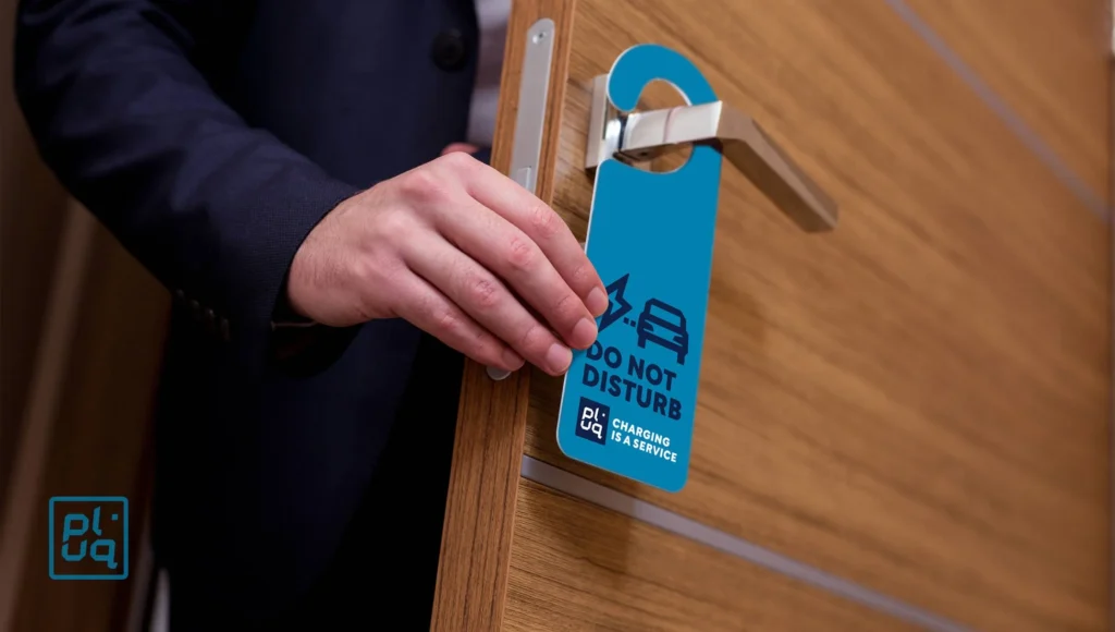 A person places a blue Do Not Disturb sign with a charging car symbol on a silver door handle of a wooden door, echoing the toekomst of electric mobility in Duitsland. The sign also reads CHARGING IN SERVICE.