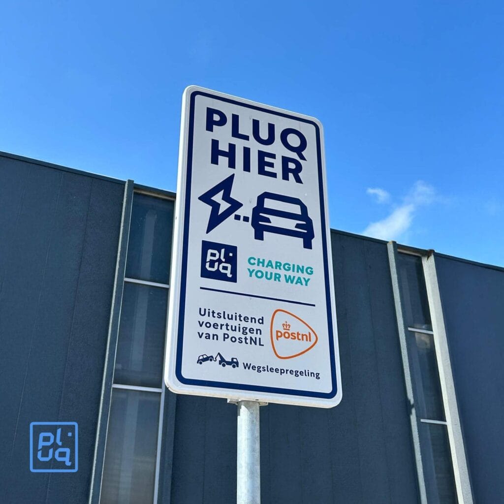 A sign against a blue sky and gray building reads “PLUQ HIER” with icons of a plug and car, marking an EV fleet charging spot reserved for PostNL vehicles at one of their dedicated hubs. PLUQ and PostNL logos are also visible.