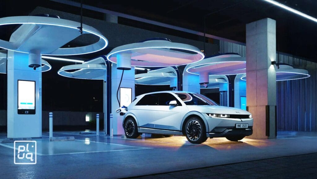 A sleek white electric car is parked at a modern charging station with futuristic blue lighting and circular overhead structures at night. Charging units and illuminated displays are visible around the car.