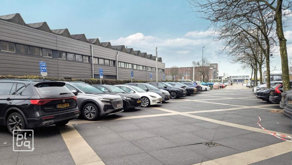 A row of cars parked in marked spaces outside a modern building with a sawtooth roof. Several parking spots are reserved for disabled drivers, indicated by blue signs. Trees line the right side of the parking area.