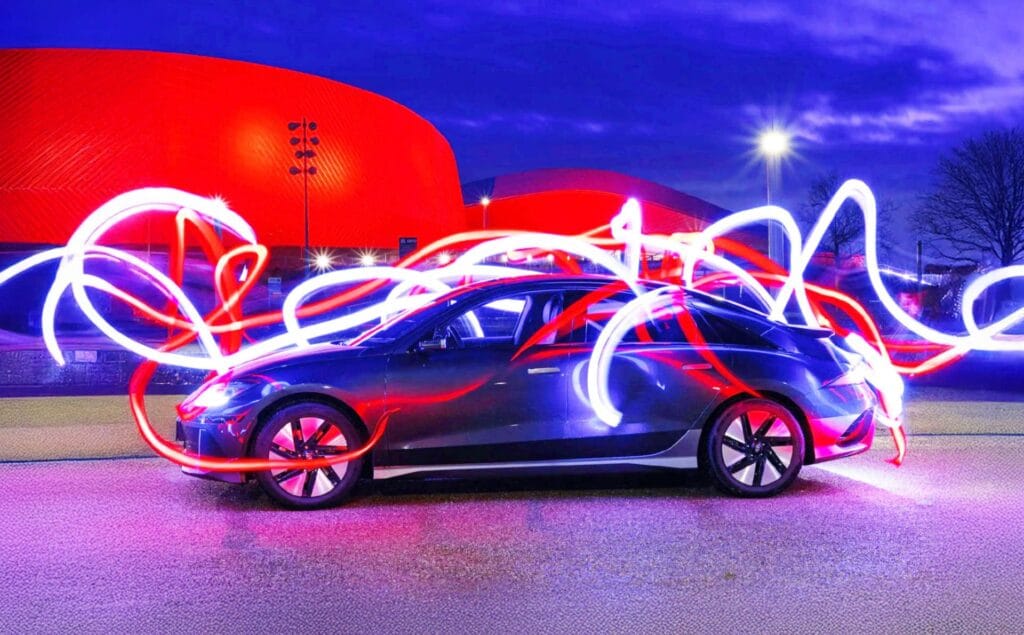 A sleek car is parked at night in front of a bright red building, with swirling red and white light trails encircling the vehicle and illuminating the scene.