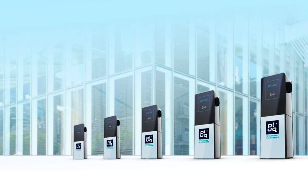 A row of five electric vehicle charging stations is lined up outdoors in front of a modern glass building, with each station displaying the pluq logo.