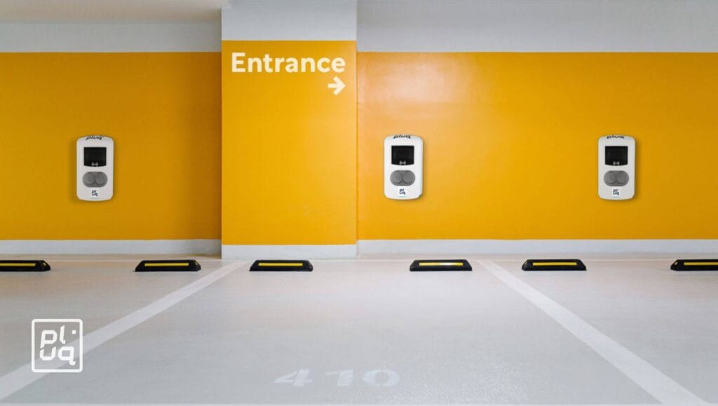 A clean, brightly lit parking area with four EV charging stations mounted on a yellow and white wall. An arrow labeled Entrance points right. Parking spots are marked on the white floor.