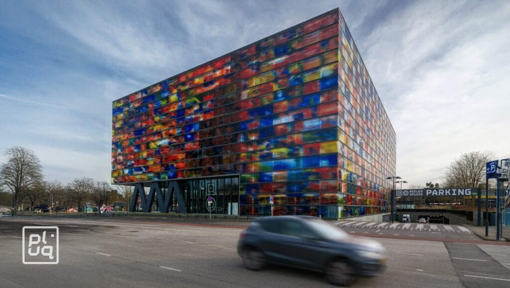 A blurred car drives past a large, colorful glass building with a checkerboard pattern. The structure has vivid, multicolored panels and a parking entrance sign is visible on the right. It’s a cloudy day.