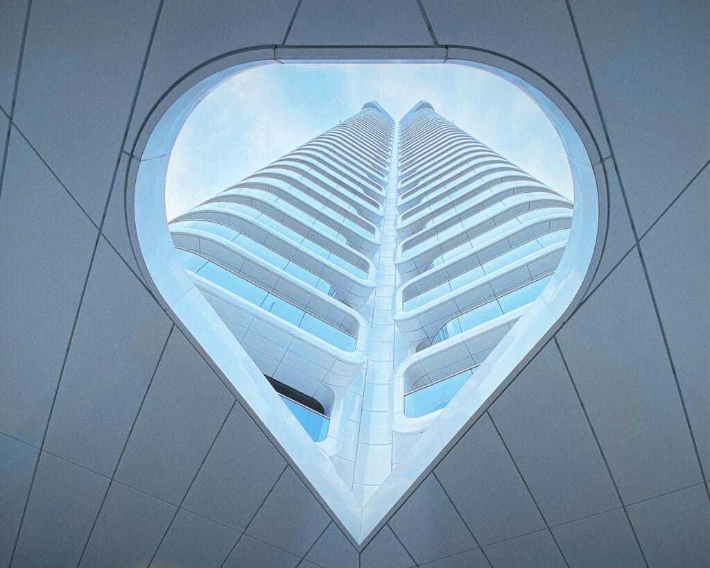 A tall, modern skyscraper is viewed from below through a large, triangular opening in the ceiling, framing the building against a blue sky with soft clouds. The building features white, curved, symmetrical lines.