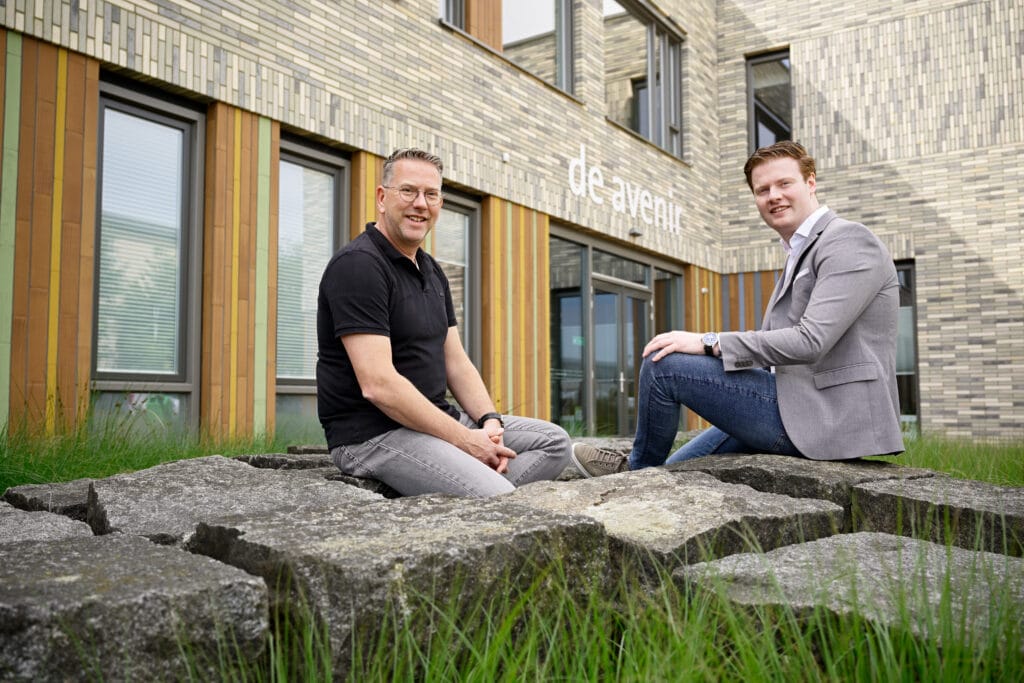 Two men sit on large stone blocks in front of a modern building with de avent written on it, surrounded by green grass. One man wears a black shirt, the other a grey blazer and jeans.