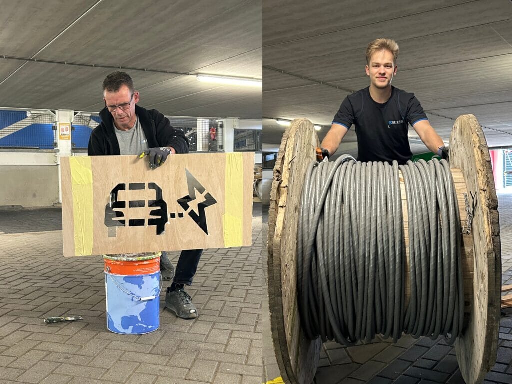 A man paints a charging station symbol on wood, while another man stands behind a large spool of thick cable in a parking garage.