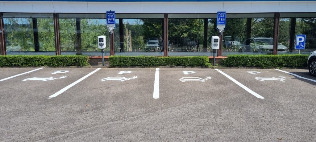 Five parking spaces for electric vehicles with charging stations in front of a building with large windows; greenery and reflections of trees visible in the background.