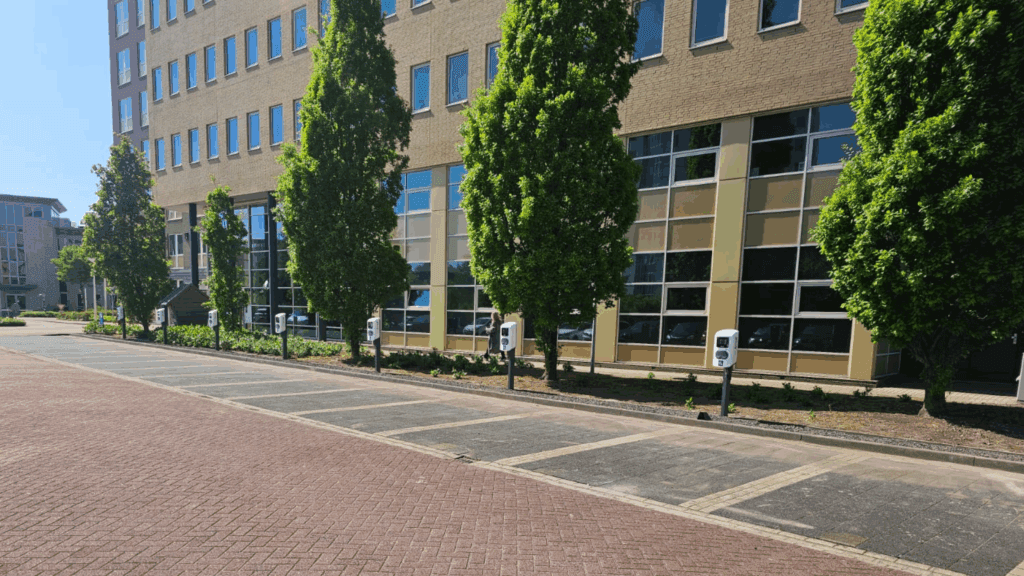 A row of empty parking spaces with electric vehicle charging stations is situated next to a modern office building lined with tall green trees on a sunny day.