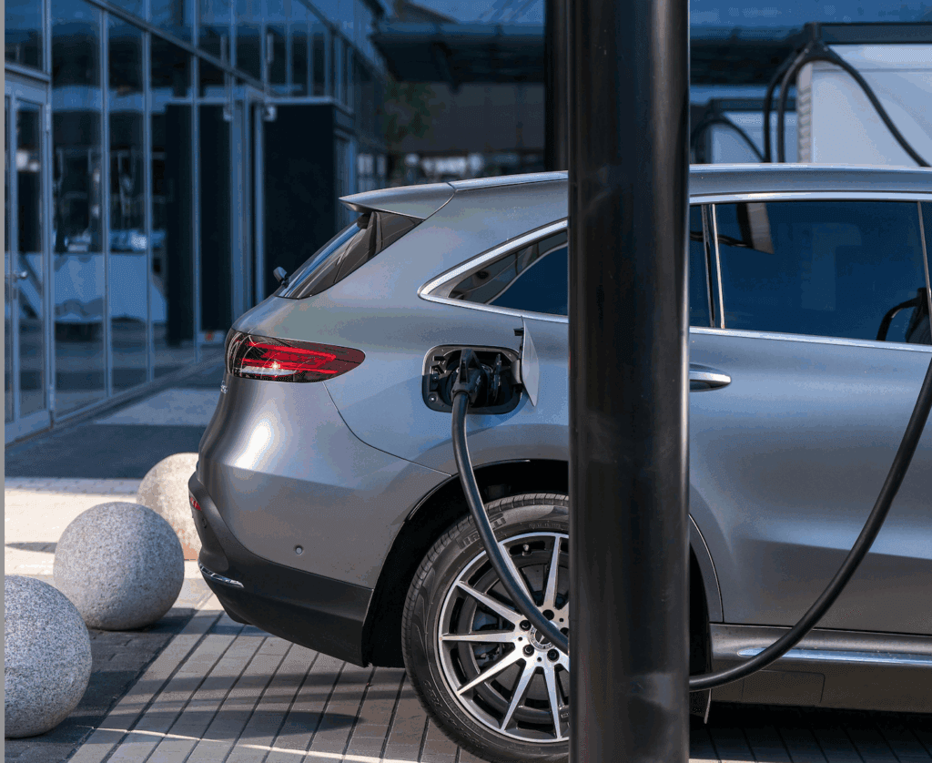 A silver electric car is parked outdoors, charging at a charging station. The vehicle is plugged in on the rear side, with modern glass buildings reflected in the background and round stone bollards nearby.