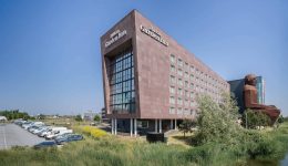 A modern, angular Hilton Garden Inn hotel with red-brown exterior walls, large glass windows, and a grassy foreground. Several cars, including electric cars at the EV charge station, are parked in a lot to the left under a clear blue sky.