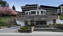 The entrance of the modern Traube Tonbach hotel features curved architecture, large windows, landscaped gardens, blooming pink trees, and convenient EV charging stations under a clear blue sky.