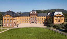 A large, yellow baroque-style palace with a central courtyard, surrounded by green lawns and trees, features discreet EV charging stations, set against a backdrop of forested hills under a clear blue sky.