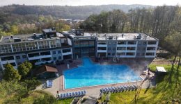 Aerial view of a large, modern hotel with outdoor pool emitting steam, surrounded by trees and greenery, and loungers around the pool. Forested hills and buildings are visible in the background, with free loading stations available to charge electric cars.