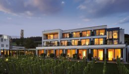Modern three-story building with large windows and balconies, warmly lit at dusk, overlooks a vineyard beneath a partly cloudy sky—plus free Pluq charging stations available for your electric vehicle.