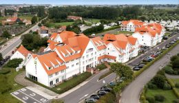 Aerial view of a large white hotel with red-orange roofs, green lawns, parked cars, and nearby houses in a suburban setting, featuring free charging stations for guests’ vehicles amid the trees and roads.