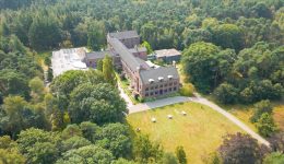 Aerial view of a large brick building surrounded by dense trees and greenery, with a manicured lawn, picnic tables, and EV charge stations in front, set against forested areas in the background.