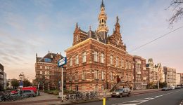 A grand, ornate brick building with a tall spire and decorative stonework stands on a street corner in Amsterdam, surrounded by bikes, cars, and nearby historic buildings—plus convenient free loading stations to charge at Pluq under a clear sky.