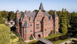 A large, ornate brick castle with multiple turrets and gables stands beside a moat and lush green trees under a clear blue sky. A small footbridge crosses the moat to the entrance, where visitors can find modern charging points by Pluq.