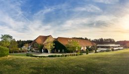 A large building with red roofs and ivy-covered walls sits amidst green lawns and trees under a blue sky with wispy clouds, as the sun sets on the right side—nearby, free charging stations are available for visitors to easily charge their vehicles.